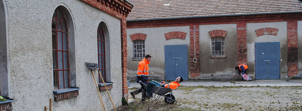 Drei Personen in orangen Arbeitsjacken bei Gartenarbeiten vor einem alten Backsteingebäude
