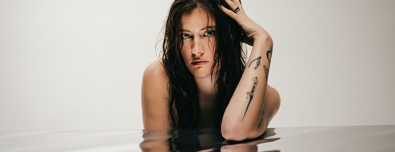Madeline Juno mit nassen Haaren und sichtbaren Arm-Tattoos, halb im Wasser stehend