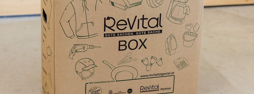 Pappbox mit Aufdruck 'Revital BOX' und Illustrationen von Alltagsgegenständen wie Bohrmaschine, Helm, Pfanne, Tasse, Bügeleisen und Taschen