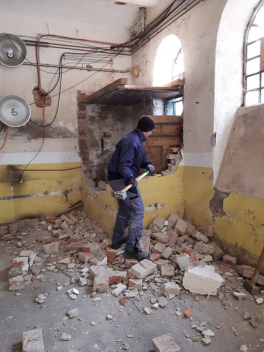 Person mit Schutzkleidung und Helm schlägt mit Vorschlaghammer auf eine teilweise eingestürzte Wand in einem Raum mit gelb-weiß gestrichenen Wänden