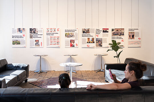 © Marija Šabanović Zwei Personen sitzen auf schwarzen Sofas in einem Raum mit weißen Wänden, an denen mehrere Informationsplakate hängen, und einem Teppich auf dem Boden.