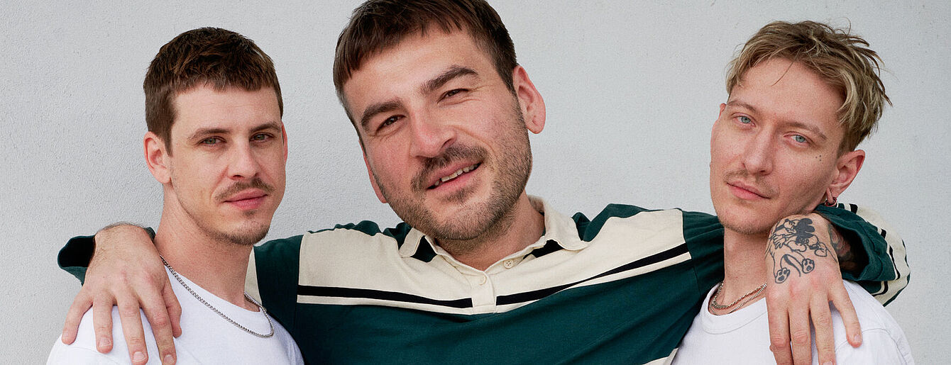 Drei Personen stehen eng zusammen vor weißer Wand, zwei tragen weiße T-Shirts, eine ein grün-weißes Hemd