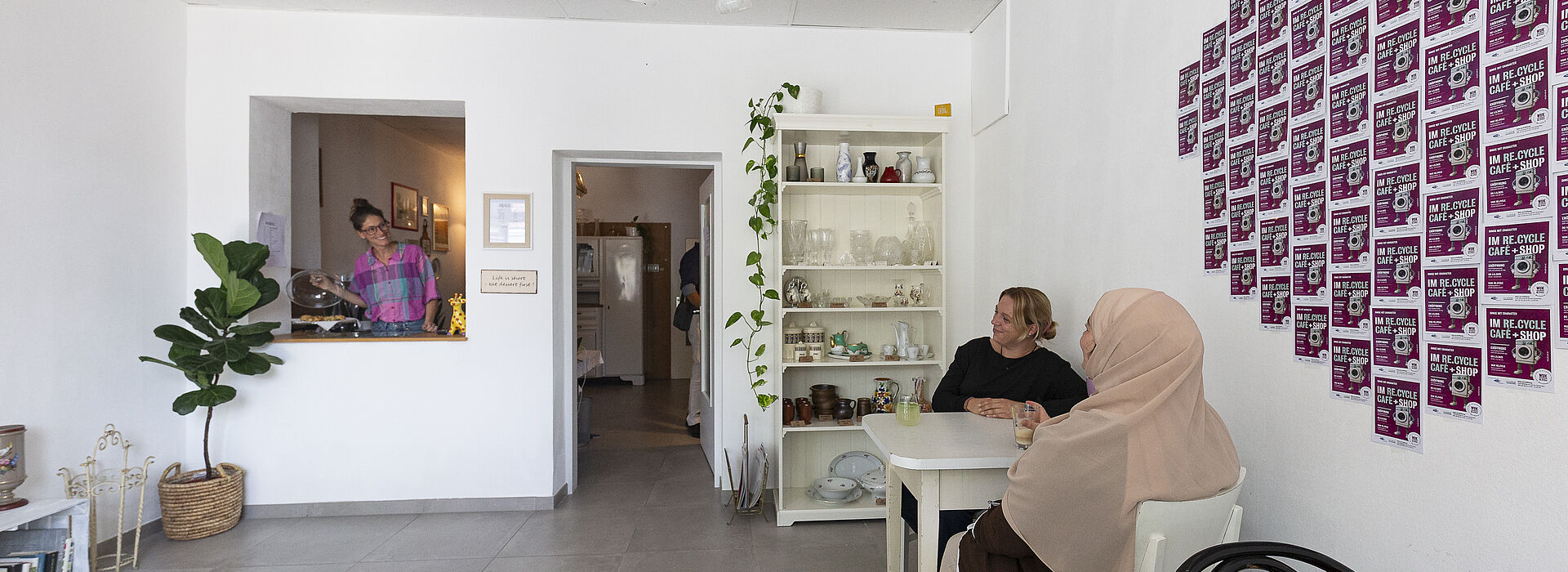 Zwei Frauen sitzen an einem weißen Tisch in einem hellen Raum mit weißen Wänden, an der Wand hängen viele kleine quadratische Bilder in einem Raster, im Hintergrund steht ein Regal mit Gläsern und Pflanzen, links ist eine Durchreiche mit einer Person dahinter.