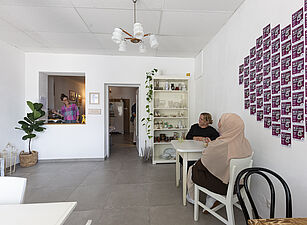 Zwei Frauen sitzen an einem weißen Tisch in einem hellen Raum mit weißen Wänden, an der Wand hängen viele kleine quadratische Bilder in einem Raster, im Hintergrund steht ein Regal mit Gläsern und Pflanzen, links ist eine Durchreiche mit einer Person dahinter.