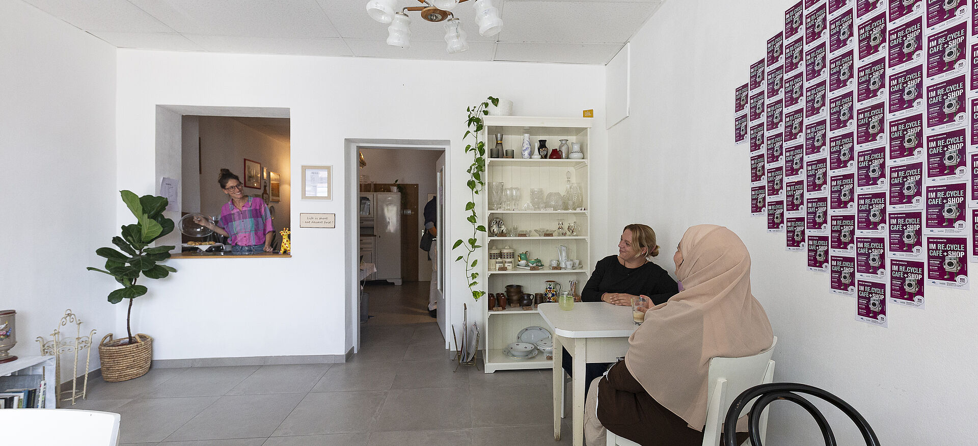 Zwei Frauen sitzen an einem weißen Tisch in einem hellen Raum mit weißen Wänden, an der Wand hängen viele kleine quadratische Bilder in einem Raster, im Hintergrund steht ein Regal mit Gläsern und Pflanzen, links ist eine Durchreiche mit einer Person dahinter.