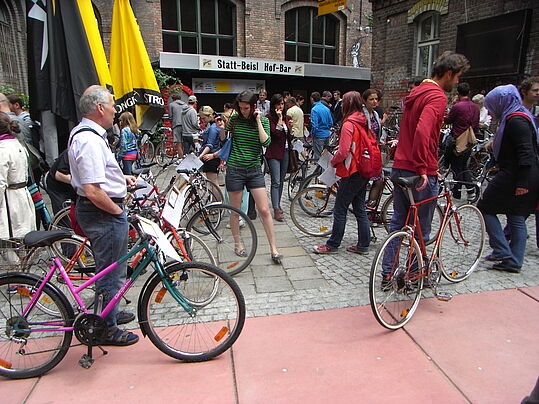 Menschen mit Fahrrädern versammeln sich vor einem Gebäude mit dem Schild 'Statt-Beisl Hof-Bar' bei einer Fahrrad Selbsthilfe Werkstatt