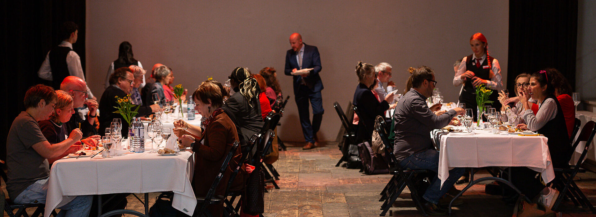 Mehrere Personen sitzen an gegenüberstehenden, weiß gedeckten Tischen und essen, im Hintergrund steht ein Mann mit Notizblock.