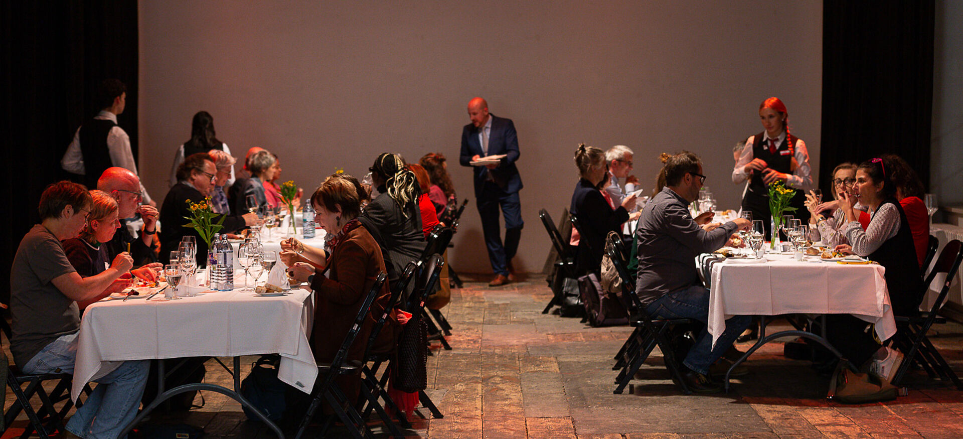 Mehrere Personen sitzen an gegenüberstehenden, weiß gedeckten Tischen und essen, im Hintergrund steht ein Mann mit Notizblock.