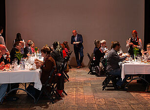 Mehrere Personen sitzen an gegenüberstehenden, weiß gedeckten Tischen und essen, im Hintergrund steht ein Mann mit Notizblock.