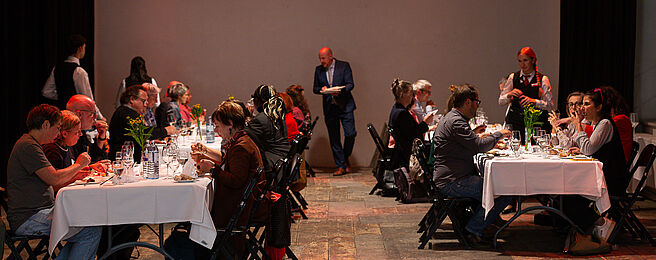 Mehrere Personen sitzen an gegenüberstehenden, weiß gedeckten Tischen und essen, im Hintergrund steht ein Mann mit Notizblock.