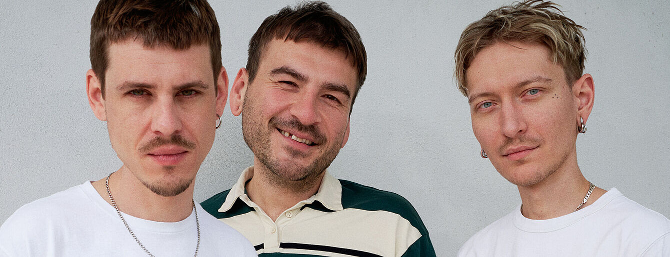 Drei Personen stehen eng zusammen vor weißer Wand, zwei tragen weiße T-Shirts, eine ein grün-weißes Hemd