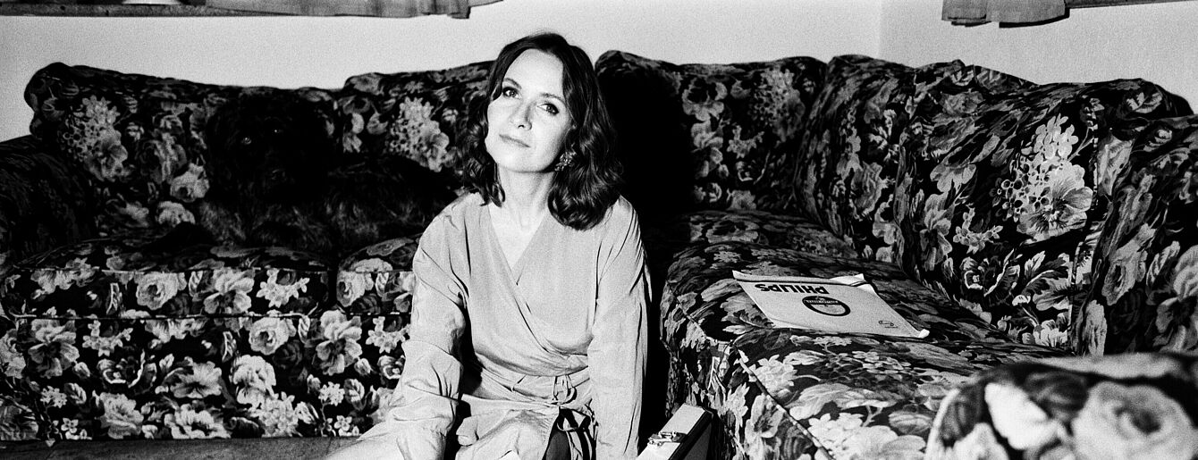 Person sitzt auf dem Boden zwischen zwei floral gemusterten Sofas, bedient einen Plattenspieler in einem Koffer, daneben ein Plattenständer mit Schallplatten.