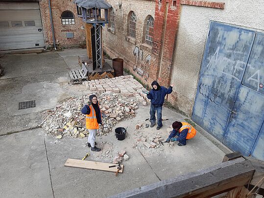 Drei Personen in Warnwesten und Arbeitskleidung reinigen und sortieren Ziegelsteine auf einem Hof mit Backsteinmauer und blauer Metalltür