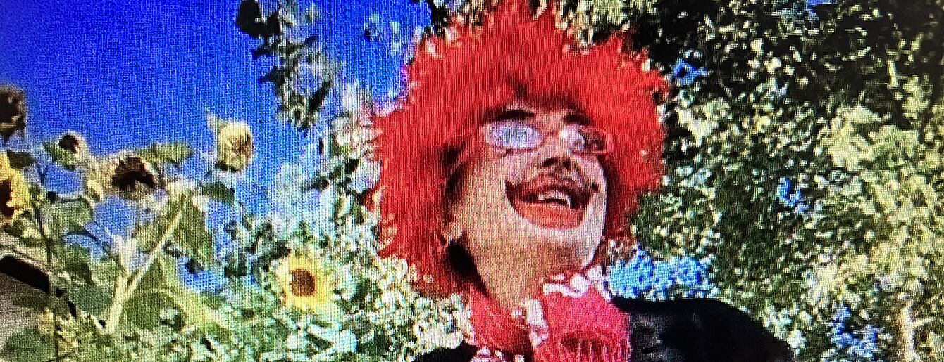 eine Clown-Person mit roten Haaren, roter Brille und rotem Ringel-Tshirt mit schwarzem Hut in der Hand vor ein paar Sonnenblumen, im Hintergrund ein Baum