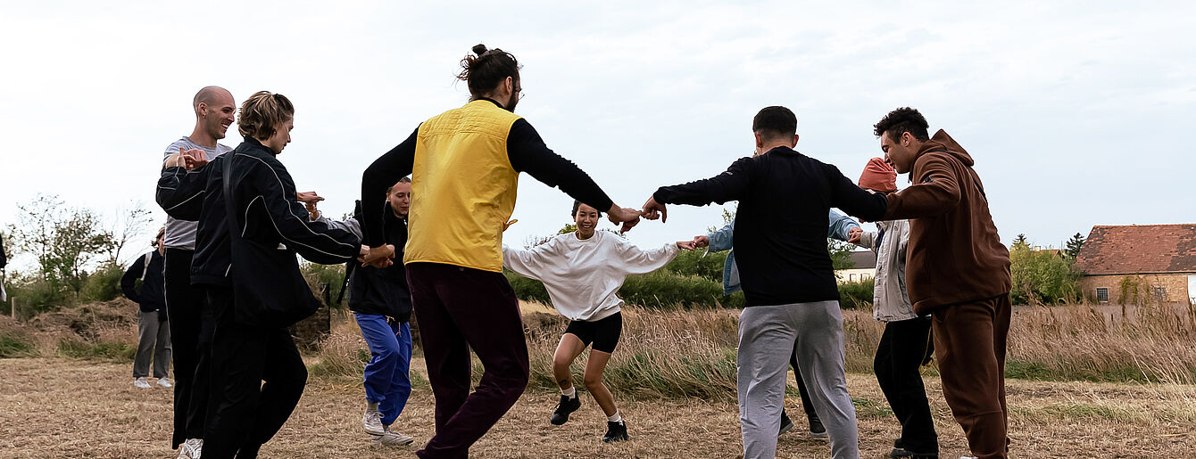 Eine Gruppe von Jugendlichen, die sich im Kreis im Freien an den Händen halten. Sie sind auf einem Feld. Im Hintergrund ist ein Haus.