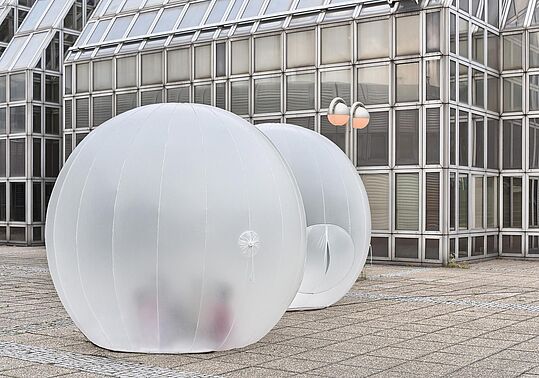 Zwei durchsichtige, aufblasbare Kugeln auf einem gepflasterten Platz vor einem modernen Glasgebäude
