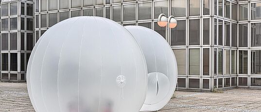 Zwei durchsichtige, aufblasbare Kugeln auf einem gepflasterten Platz vor einem modernen Glasgebäude
