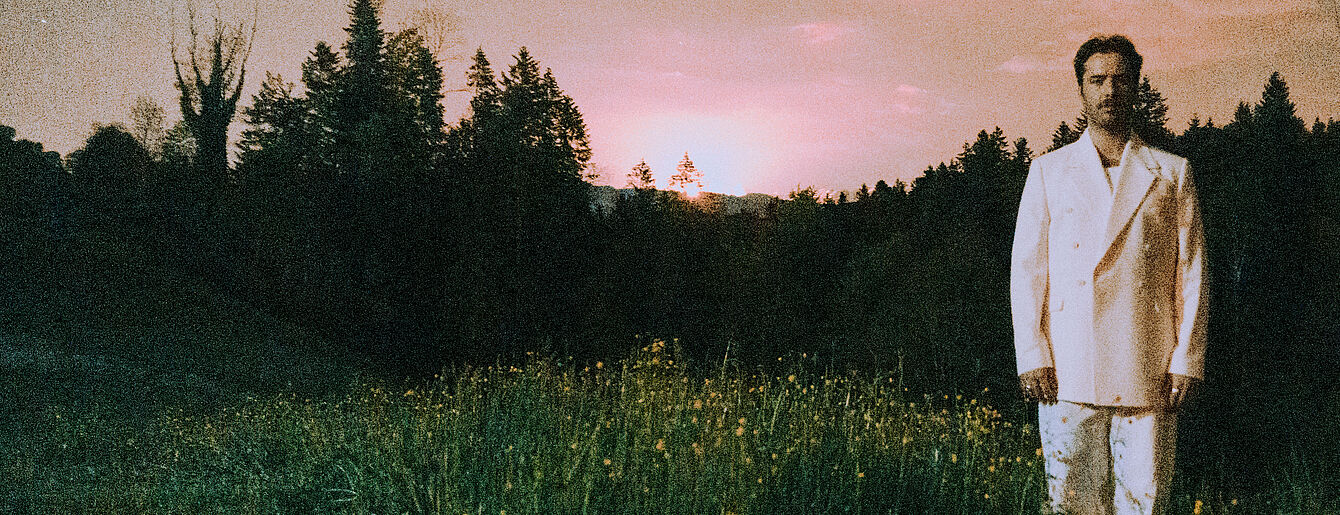 Benjamin Amaru steht in einem weißen Anzug auf einer Wiese vor einem Wald bei Sonnenuntergang