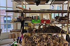 Eine Konstruktion aus Holzregalen steht in einem Raum mit großen Fenstern. Die Regale sind aus verschiedenen Holzarten und -farben, einige Bretter sind horizontal, andere vertikal angeordnet.