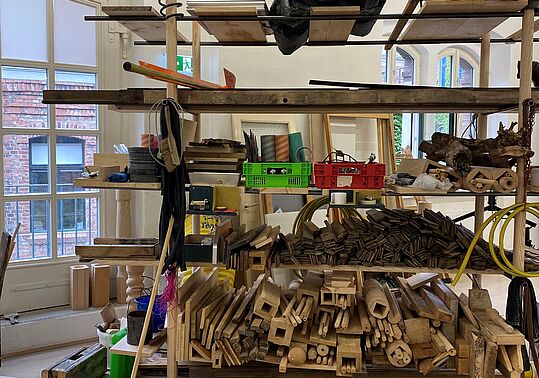 Eine Konstruktion aus Holzregalen steht in einem Raum mit großen Fenstern. Die Regale sind aus verschiedenen Holzarten und -farben, einige Bretter sind horizontal, andere vertikal angeordnet.