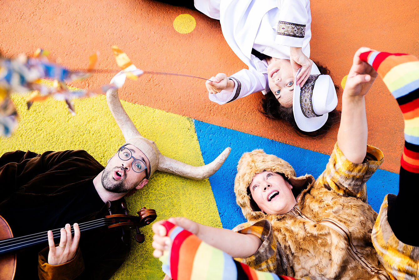 Drei Personen liegen auf einem bunten Teppich, eine spielt Geige, eine hält ein großes Auerochsengeweih, eine trägt ein bunt gestreiftes Kleidungsstück.