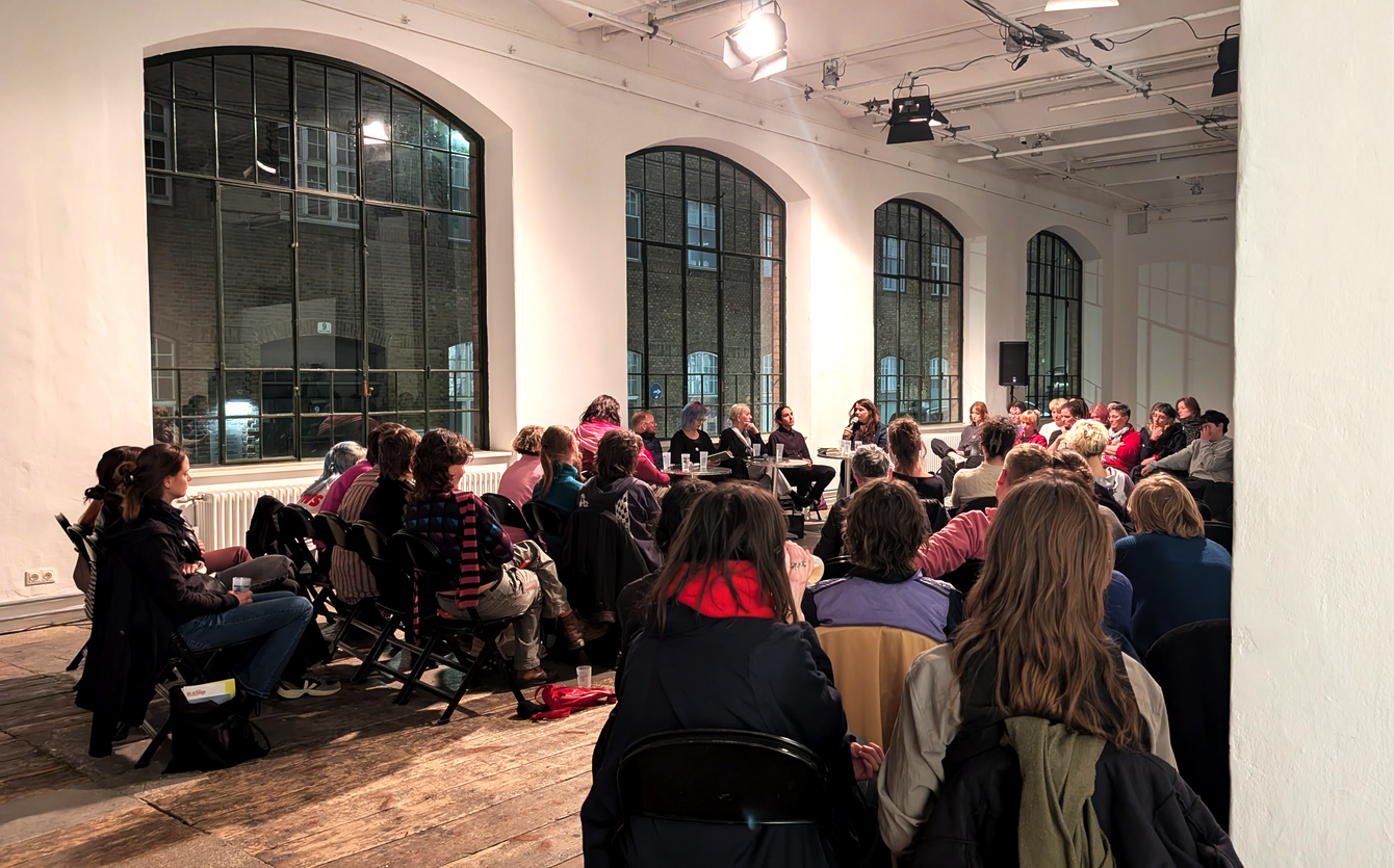 Gruppe von Menschen sitzt in einem großen Raum mit hohen Fenstern und hört einer Diskussionsrunde zu