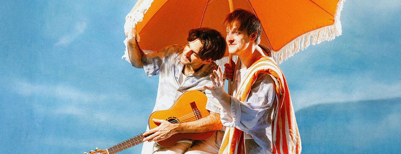 Zwei junge Personen sitzen unter einem orangefarbenen Sonnenschirm, einer hält eine Gitarre, der andere trägt ein gestreiftes Handtuch über der Schulter
