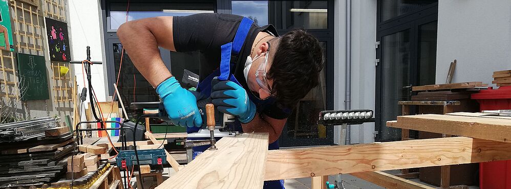 Person mit Schutzbrille und Handschuhen arbeitet an Holzbrett in Werkstatt