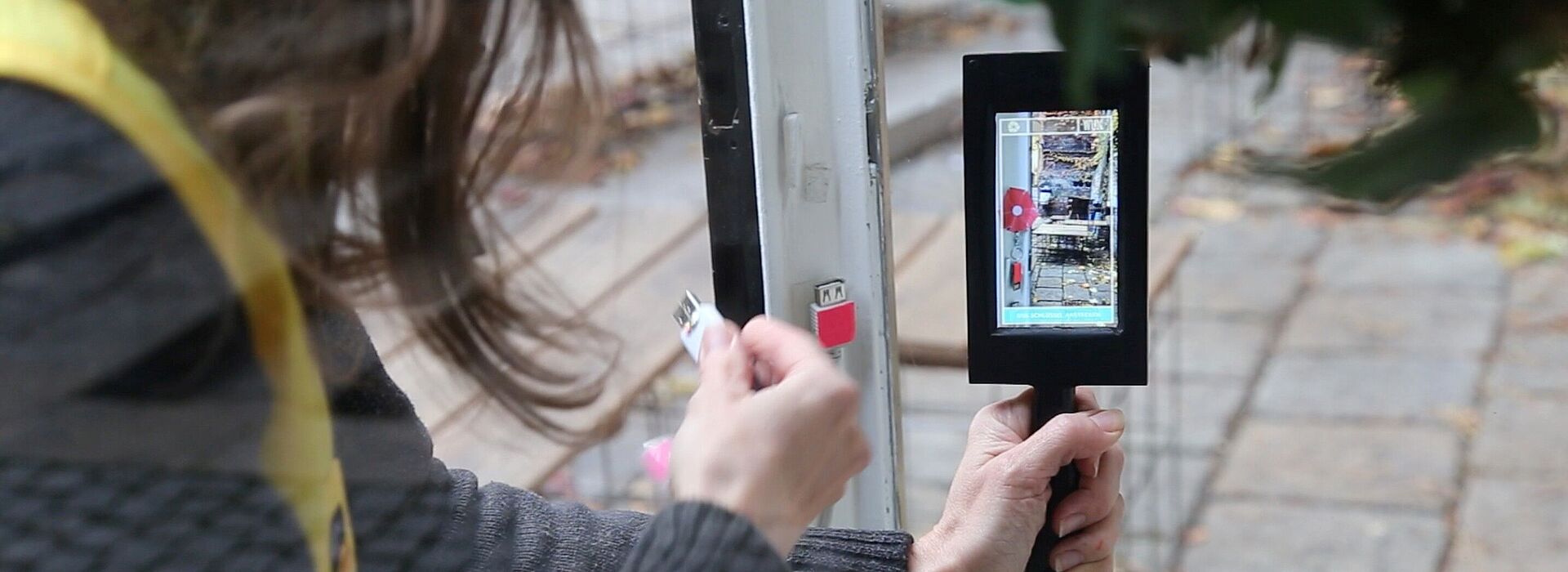 Person hält ein Smartphone mit grünem Band und steckt ein Kabel in einen Anschluss an einer Tür ein