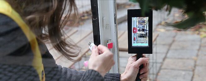 Person hält ein Smartphone mit grünem Band und steckt ein Kabel in einen Anschluss an einer Tür ein