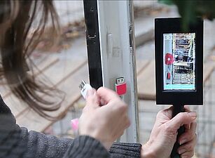 Person hält ein Smartphone mit grünem Band und steckt ein Kabel in einen Anschluss an einer Tür ein