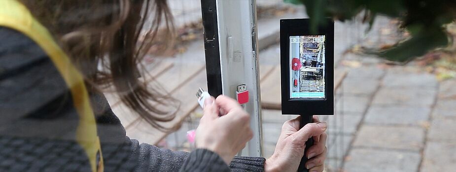 Person hält ein Smartphone mit grünem Band und steckt ein Kabel in einen Anschluss an einer Tür ein