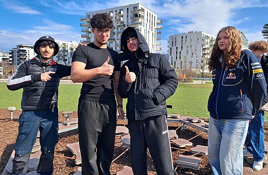 Vier Personen stehen auf einem Spielplatz. Zwei Personen zeigen Daumen hoch, eine trägt einen schwarzen Kapuzenpullover und eine andere einen schwarzen Anorak. Im Hintergrund sind moderne Wohngebäude und ein blauer Himmel.
