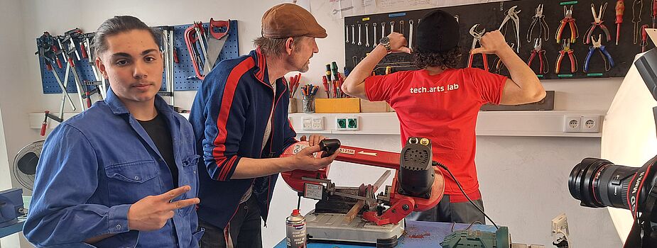 Drei Personen in einer Werkstatt. Eine Person trägt ein rotes T-Shirt mit 'tech.arts lab', eine andere hält ein Werkzeug, die dritte zeigt ein Peace-Zeichen. Auf dem Tisch liegen Werkzeuge und Materialien.
