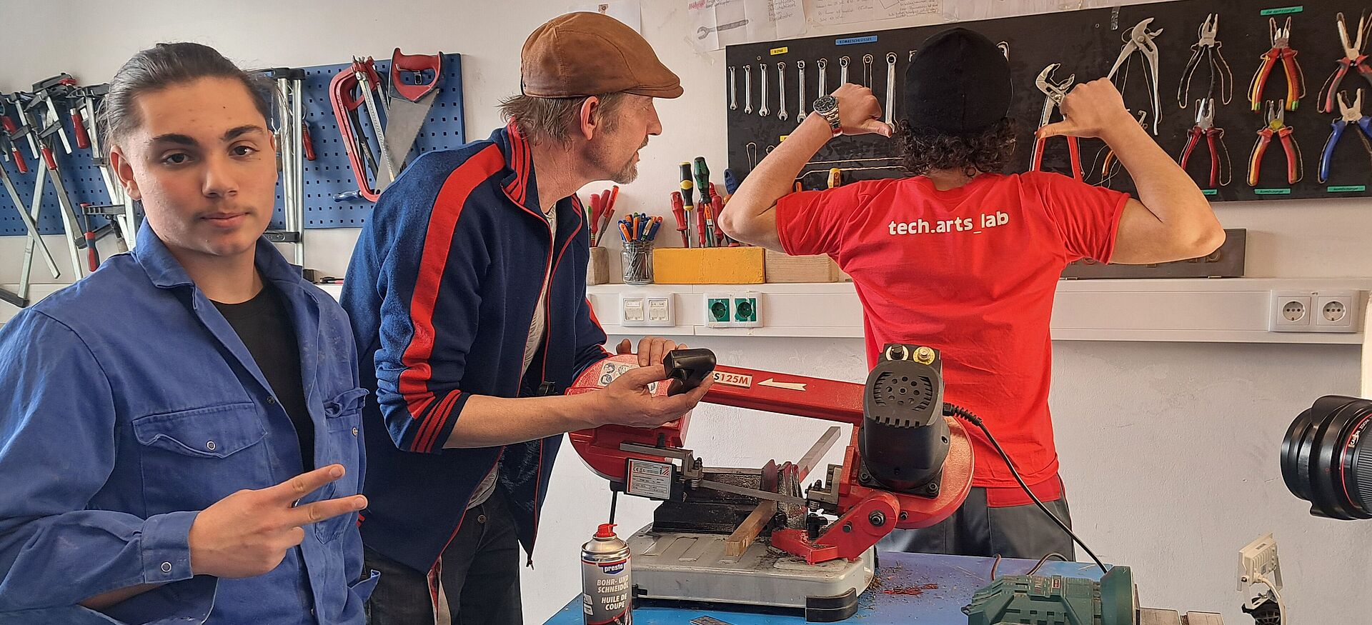 Drei Personen in einer Werkstatt. Eine Person trägt ein rotes T-Shirt mit 'tech.arts lab', eine andere hält ein Werkzeug, die dritte zeigt ein Peace-Zeichen. Auf dem Tisch liegen Werkzeuge und Materialien.