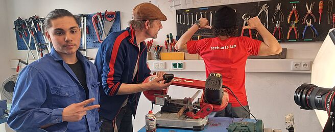 Drei Personen in einer Werkstatt. Eine Person trägt ein rotes T-Shirt mit 'tech.arts lab', eine andere hält ein Werkzeug, die dritte zeigt ein Peace-Zeichen. Auf dem Tisch liegen Werkzeuge und Materialien.
