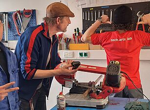 Drei Personen in einer Werkstatt. Eine Person trägt ein rotes T-Shirt mit 'tech.arts lab', eine andere hält ein Werkzeug, die dritte zeigt ein Peace-Zeichen. Auf dem Tisch liegen Werkzeuge und Materialien.