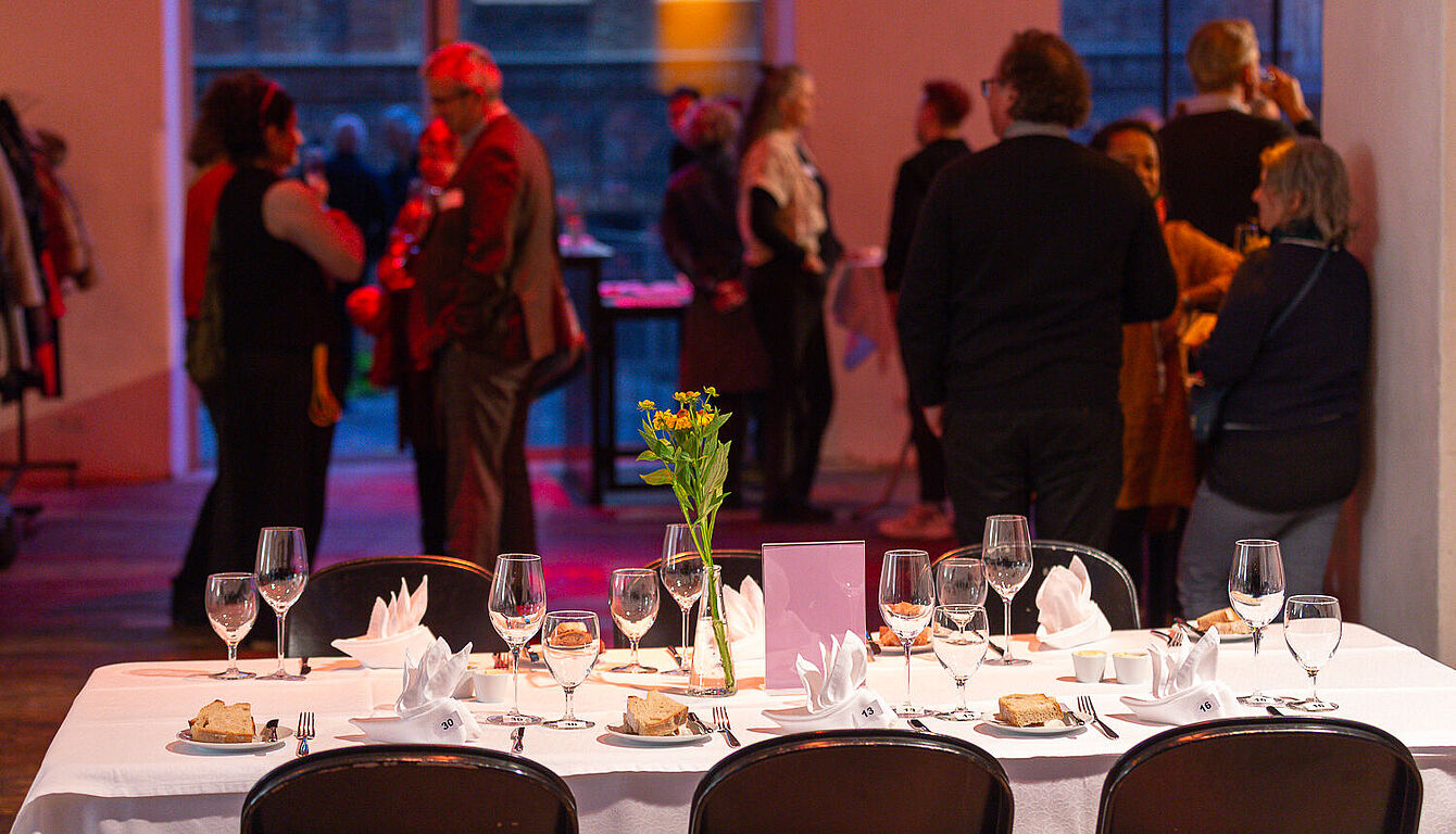 Foto: Susanne Senekowitsch Festlich gedeckter Tisch mit Gläsern, Servietten und Blumen in einem Raum, in dem mehrere Personen im Hintergrund stehen und sich unterhalten.