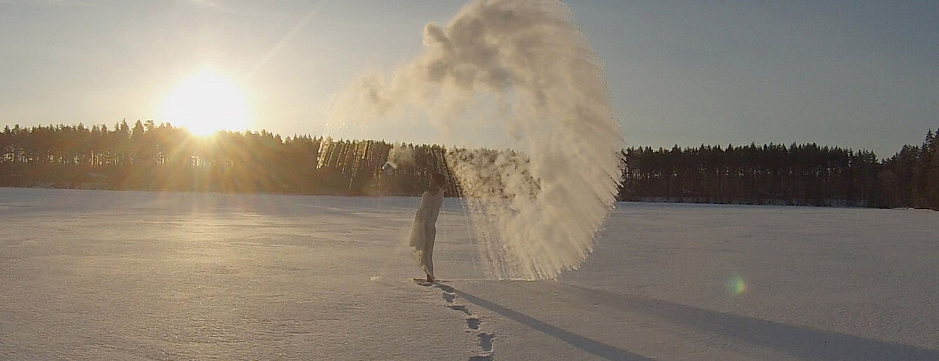 Person wirft Wasser in die Luft auf einer schneebedeckten Fläche bei Sonnenaufgang oder Sonnenuntergang mit sichtbarem Atemnebel