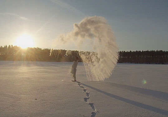 Person wirft Wasser in die Luft auf einer schneebedeckten Fläche bei Sonnenaufgang oder Sonnenuntergang mit sichtbarem Atemnebel
