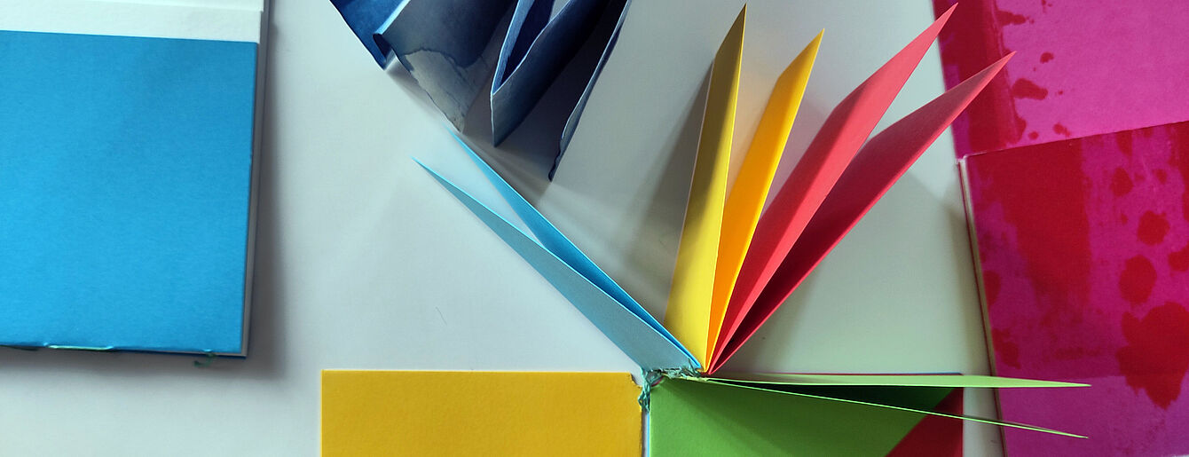 Mehrere bunte, gefaltete Papierstücke und ein aufgeschlagenes, farbiges Heft mit sichtbarer Fadenheftung auf weißem Untergrund