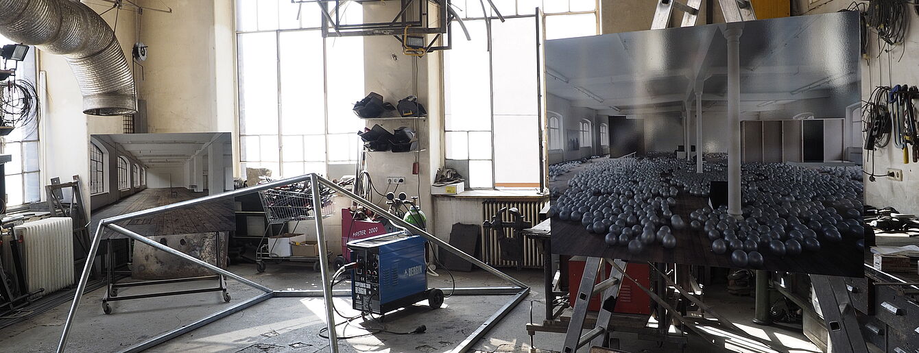 Werkstatt mit Metallrahmen, Schweißgerät und zwei großen Fotos von Innenräumen mit Säulen und Kugeln