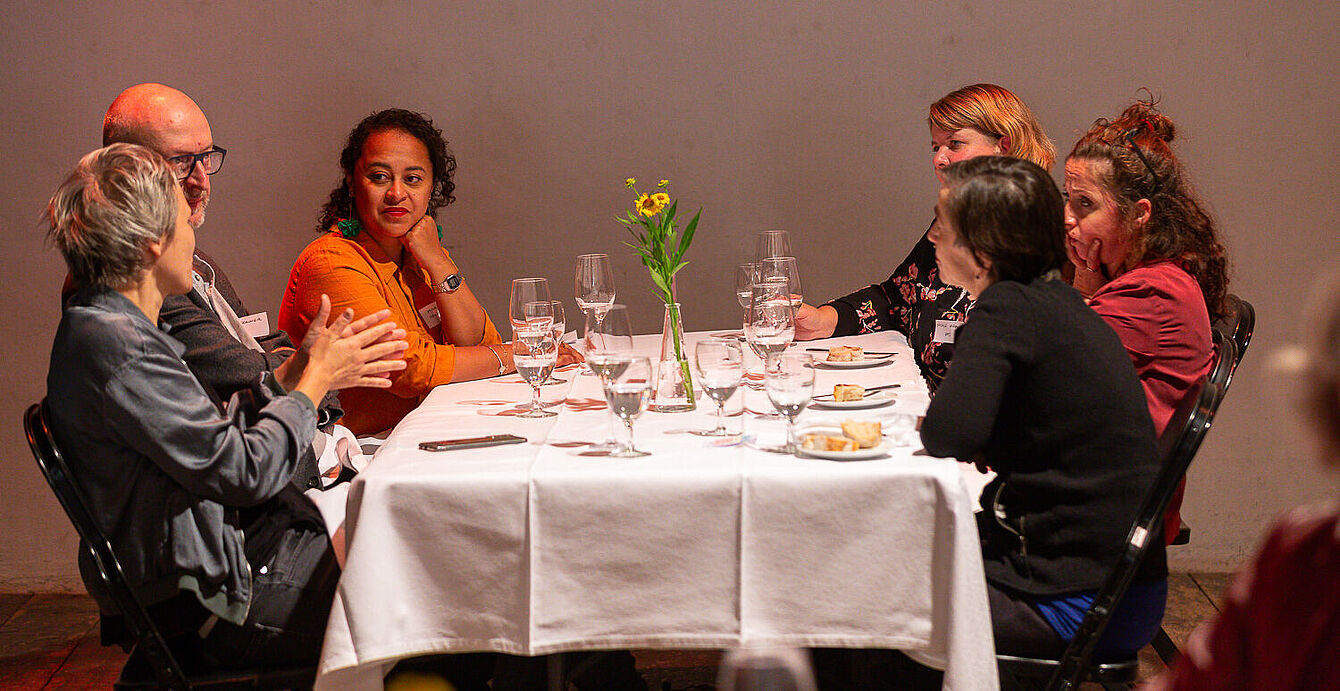 Foto: Susanne Senekowitsch Sechs Personen sitzen an einem rechteckigen Tisch mit weißer Tischdecke, darauf Gläser, Teller und eine Vase mit gelben Blumen.