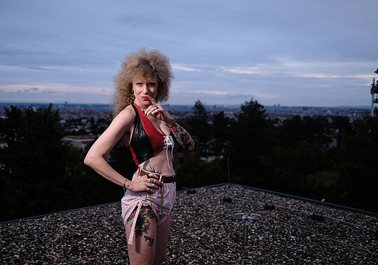 Frau mit lockigem Haar und Tattoos trägt schwarzes Top und rosa Rock und steht auf einem Dach vor einer Stadtlandschaft bei Dämmerung