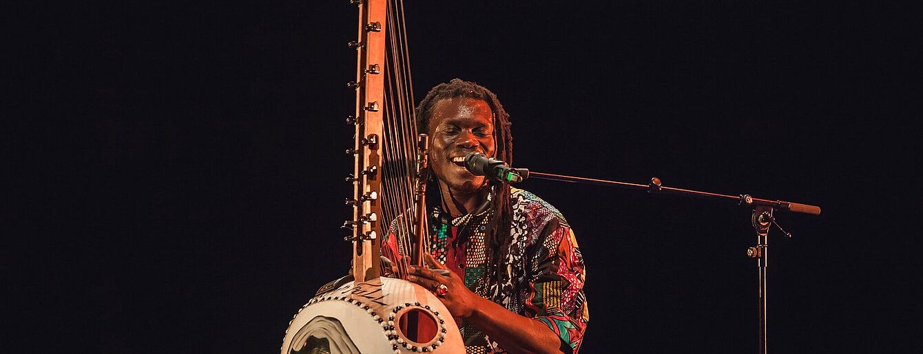 ein afrikanischer Mann spoelt auf einem saiteninstrument und singt in ein Mikrofon.