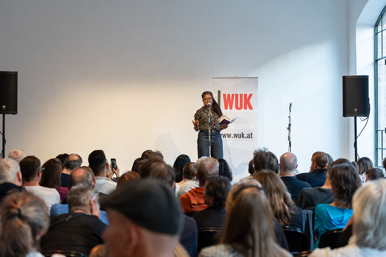 Person mit Mikrofon und Buch vor Publikum in einem hellen Raum mit WUK-Banner