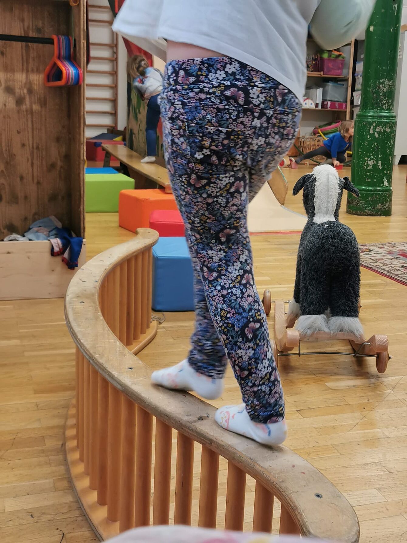 Kind mit gemusterten Hosen balanciert barfuß auf einem geschwungenen Holzgeländer in einem Spielzimmer mit Spielzeug und Teppich