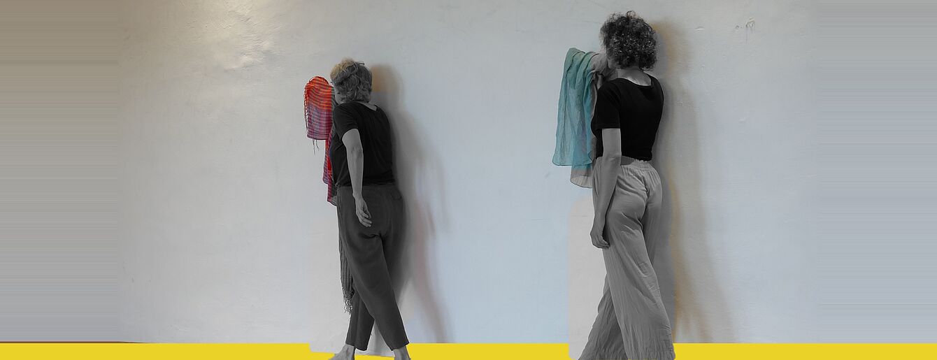 Zwei Frauen mit lockigem Haar stehen barfuß vor einer weißen Wand, jede hält ein Handtuch, eine rote und eine blaue, auf gelbem Boden