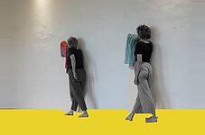 Zwei Frauen mit lockigem Haar stehen barfuß vor einer weißen Wand, jede hält ein Handtuch, eine rote und eine blaue, auf gelbem Boden