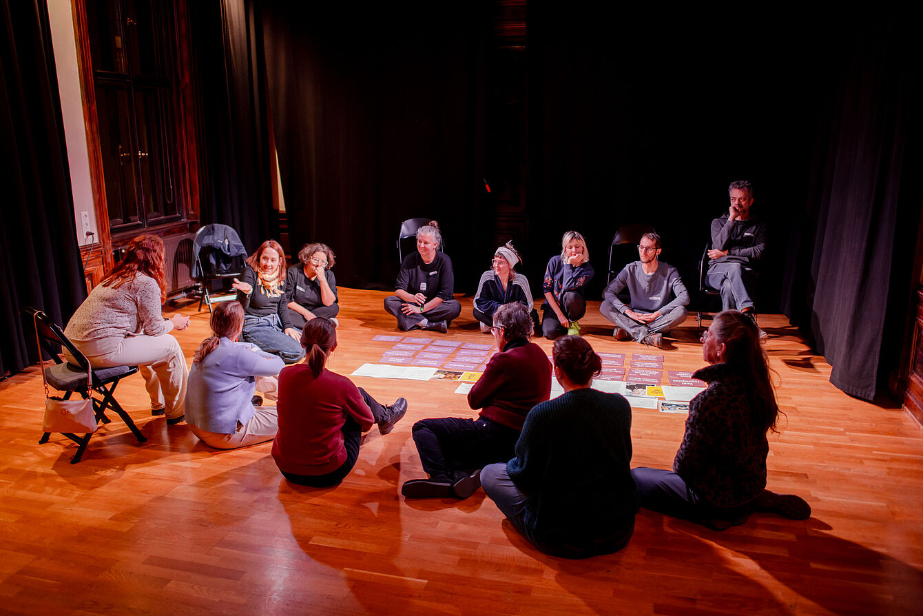 Gruppe von Menschen sitzt im Kreis auf einem Holzboden, in der Mitte liegen mehrere bunte Papiere.